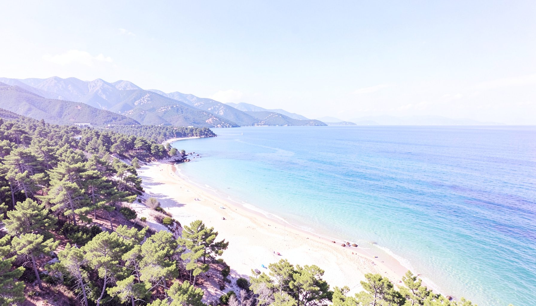 Vue aérienne d'une baie côtière avec plage de sable, mer turquoise, pins et montagnes sous un ciel bleu éclatant