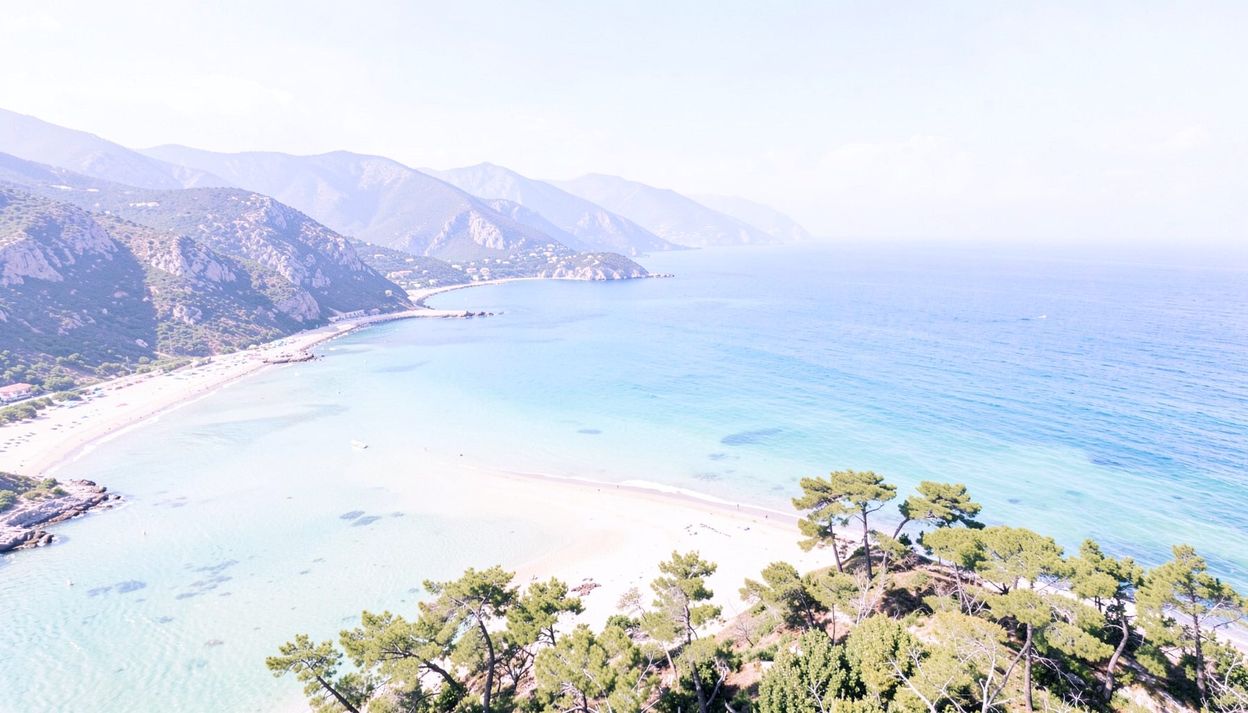 Vue aérienne d'une baie côtière avec plage de sable, mer turquoise, pins et montagnes sous un ciel bleu