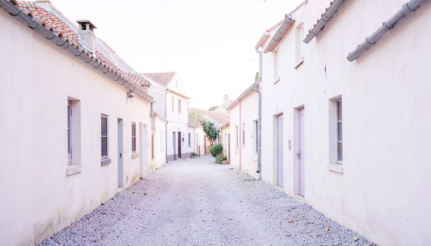 Allée de gravier bordée de bâtiments en stuc aux toits de tuiles, sous une lumière douce et naturelle