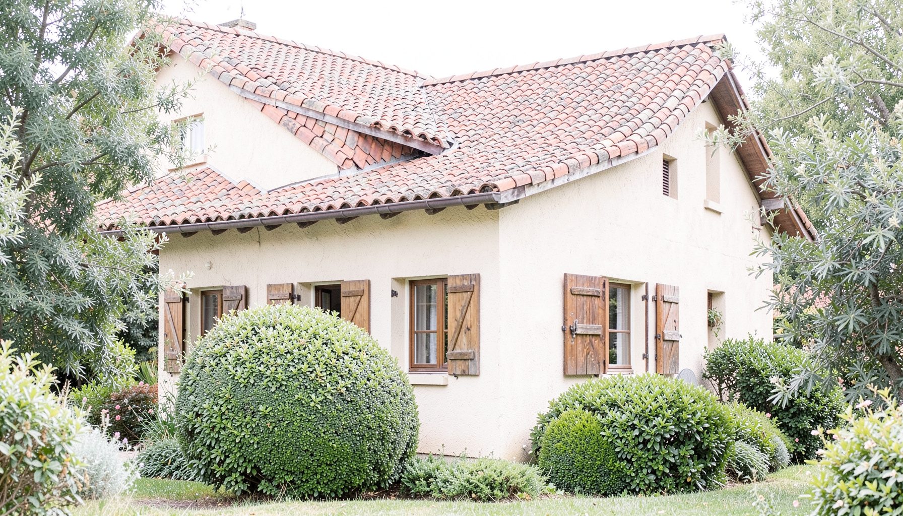 Bâtiment en stuc beige avec toit en tuiles terre cuite, volets en bois et buisson taillé en boule, entouré de verdure