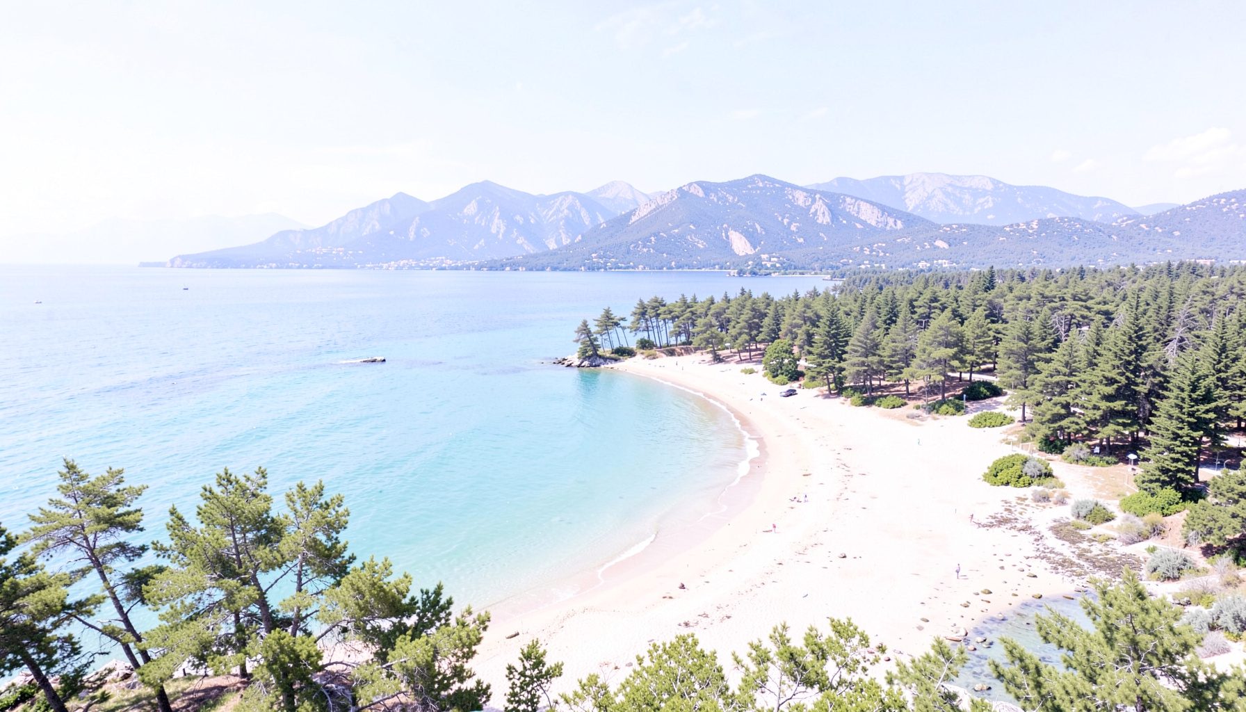 Vue aérienne d'une baie côtière avec plage de sable, mer turquoise, pins et montagnes sous un ciel bleu lumineux