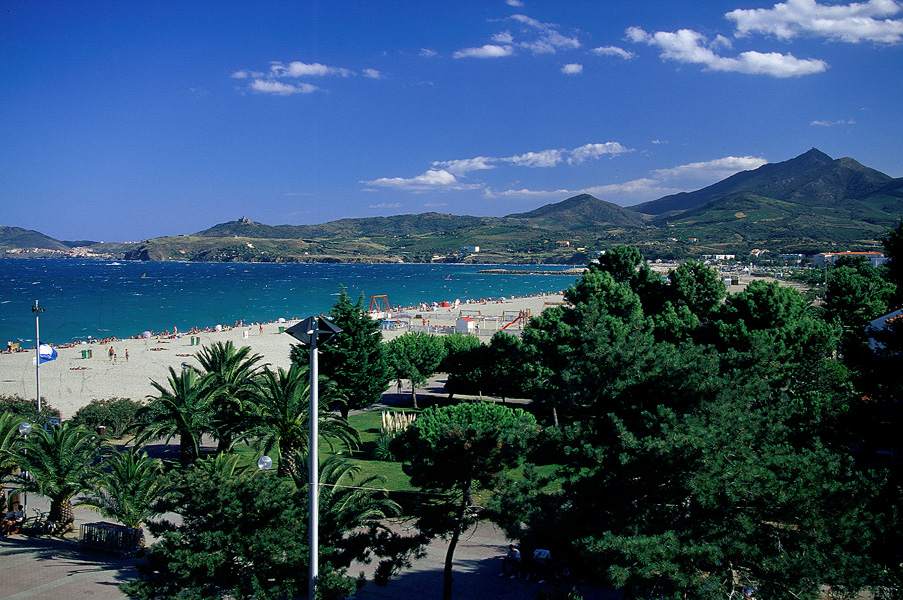 Vue aérienne d'une baie côtière avec plage de sable, mer turquoise, pins et montagnes sous un ciel bleu éclatant