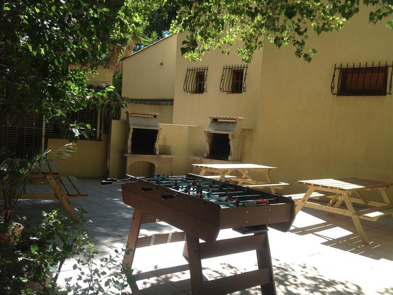 Terrasse extérieure ensoleillée avec tables de pique-nique, barbecue intégré et bâtiment aux tons jaunes et méditerranéens